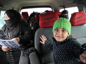 A young girl wearing a knitted green hat sits inside a minibus with women passengers sitting in the seats behind her.