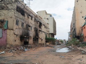 A dirt road with damaged buildings on both sides and building debris along the side of the road