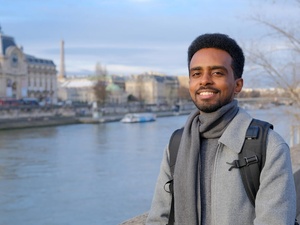 A man wearing a grey overcoat and backpack stands on a riverside with historic buildings on the opposite bank 