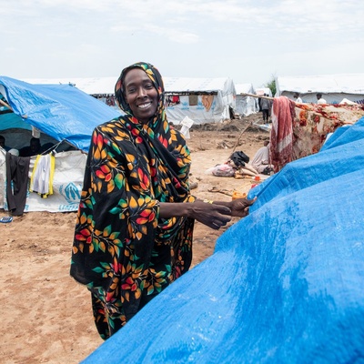 A woman smiles as she pulls a blue tarpaulin into place over a makeshift shelter.