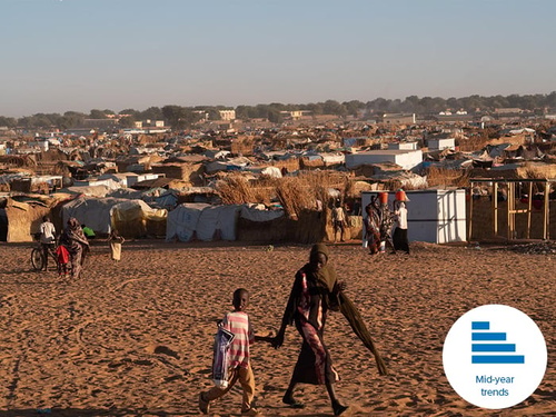 A woman holds a child's hand as they walk past a refugee tent settlement in a dusty field.