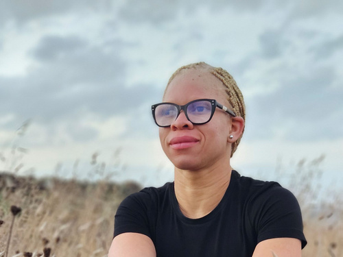 An Albino woman wearing glasses sits in a field looking into the distance, with her hair tied back