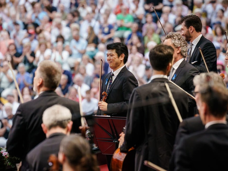 Musicians part of an orchestra stand up holding their instruments, in front of a crowd.