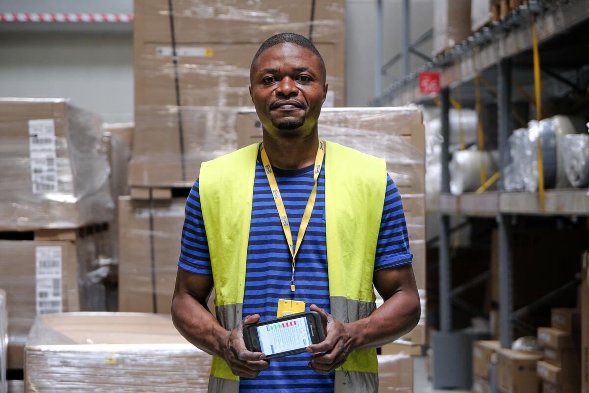 A man wearing an IKEA lanyard stands in a warehouse, holding a mobile device and smiling at the camera.
