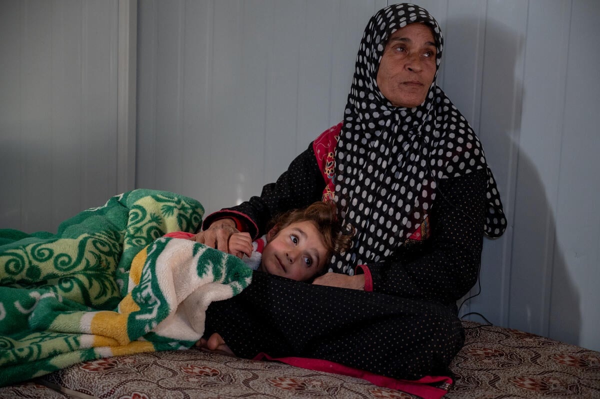 A woman sits in her shelter as her five-year-old granddaughter sleeps on her lap, covered with a blanket.
