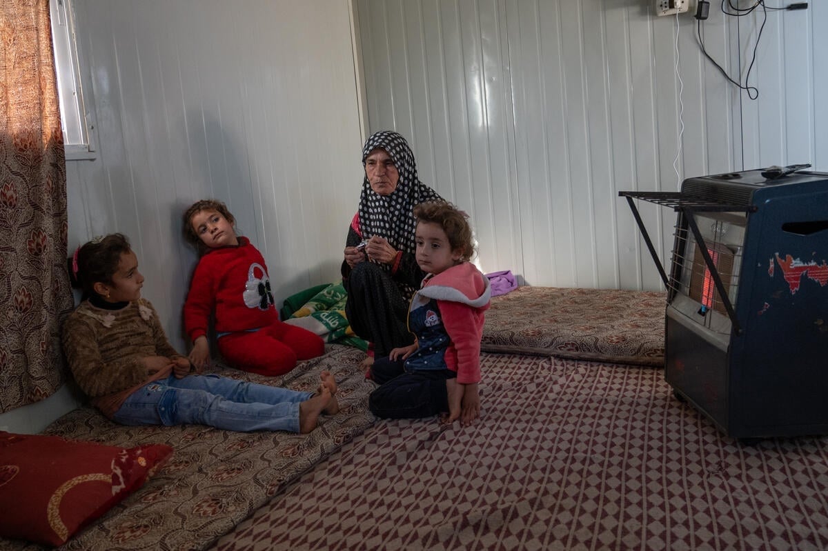 A woman sits with her grandchildren in front of a gas heater inside her shelter.