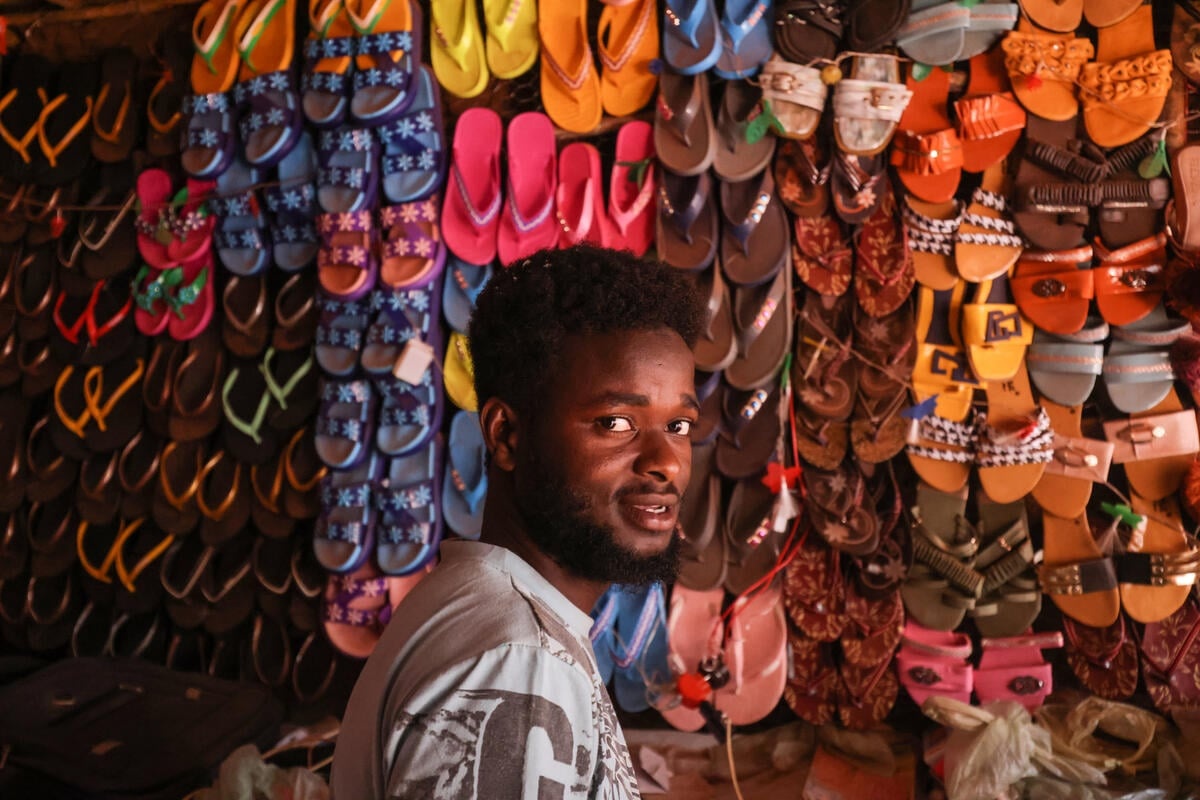A Sudanese man in front of a wall of rows of colorful sandals