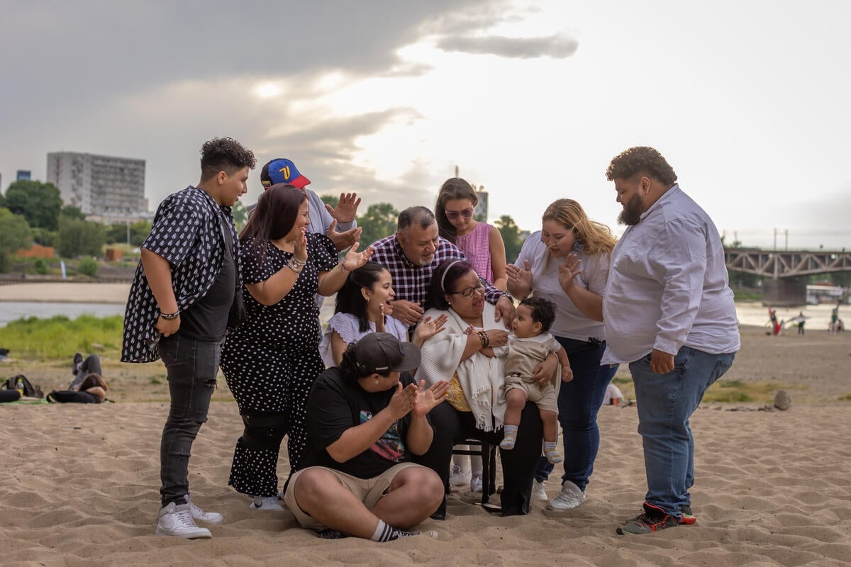 A seated woman is surrounded by family on the sandy banks of a river. A bridge, large building and a park can be seen in the background.