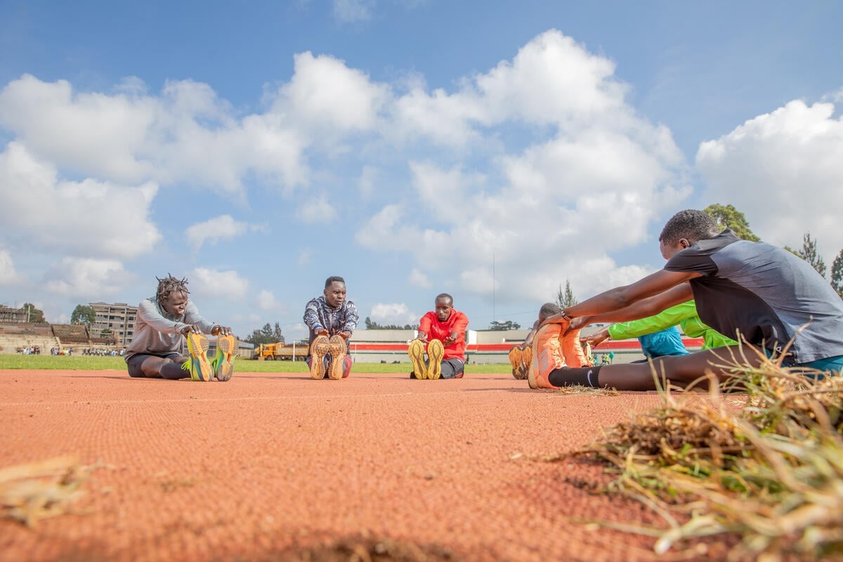 A group of athletes sit in a semi-circle on the ground of a running track touching their toes.