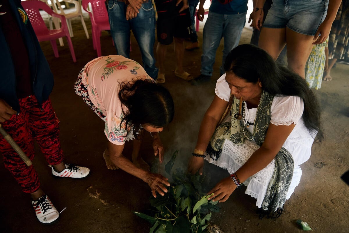 Two women crouch down over a burning pile of green leaves surrounded by onlookers.
