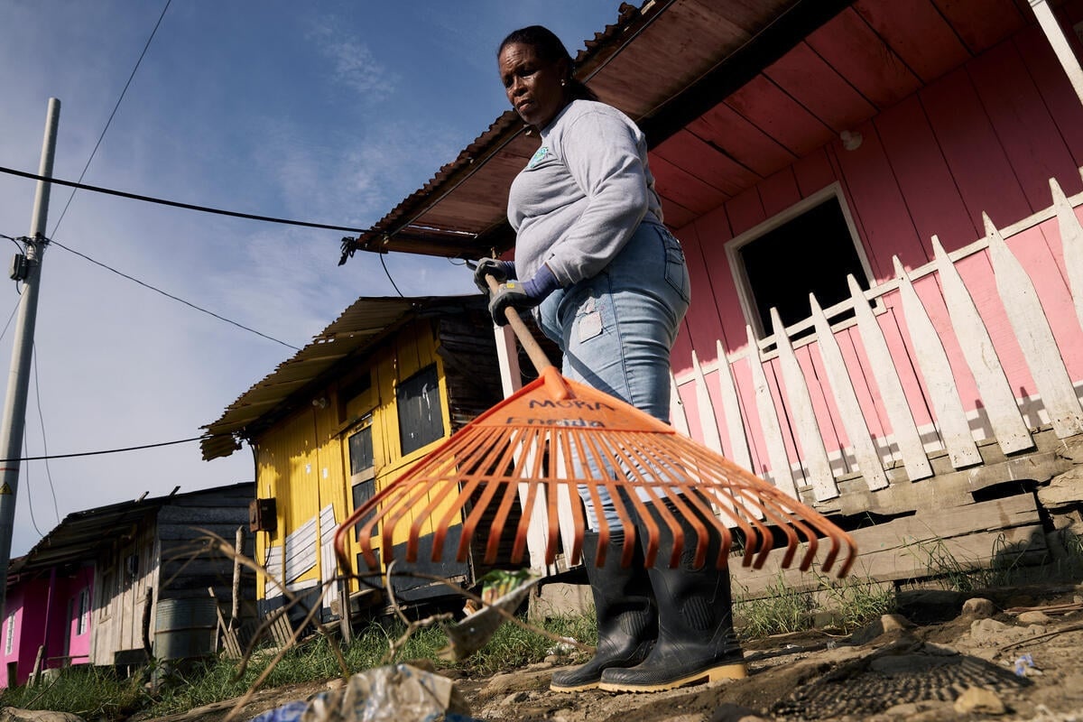A woman in rubber boots wields a rake outside a small wooden house.
