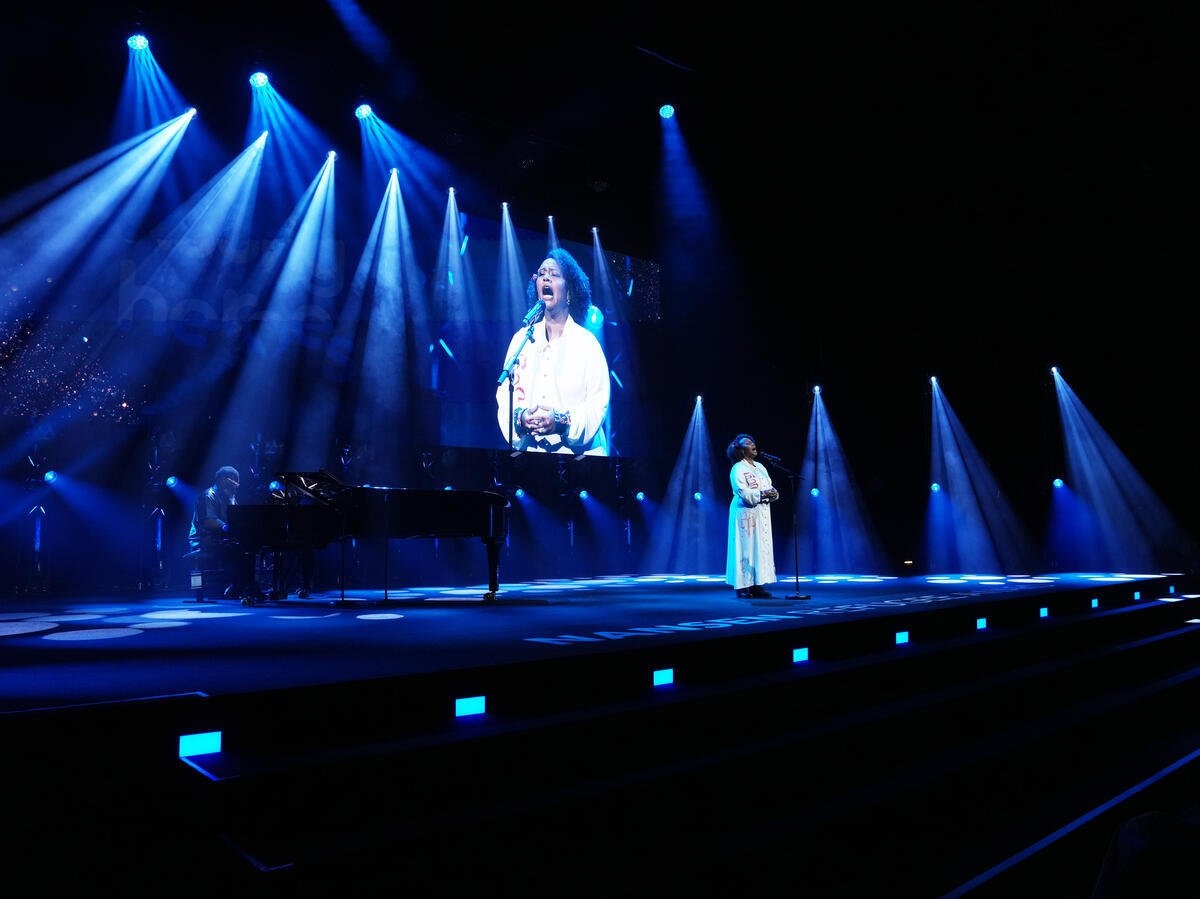 A woman in a white dress sings onstage backed by a pianist and a giant screen