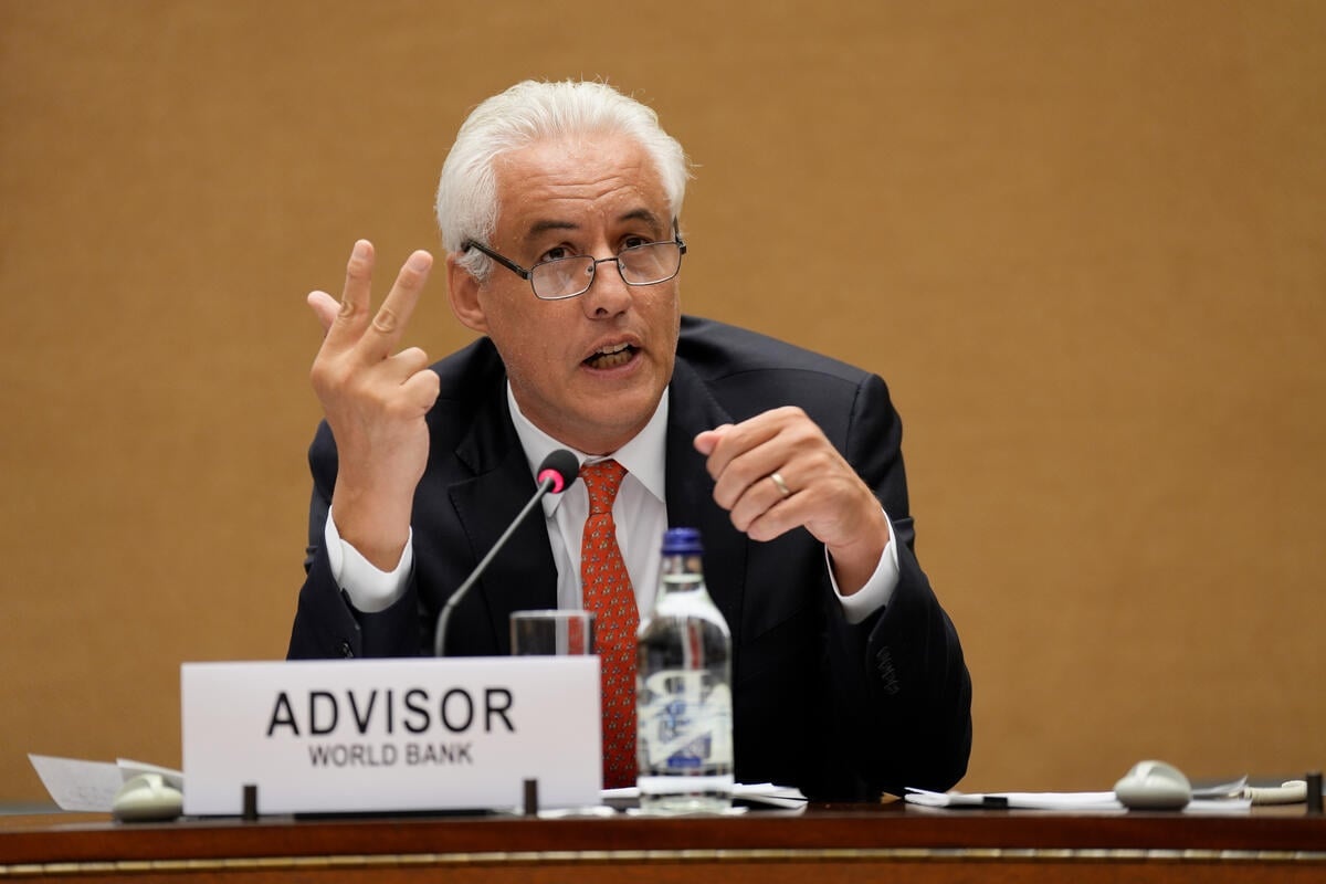 A seated man speaks into a microphone as he counts off points on his fingers