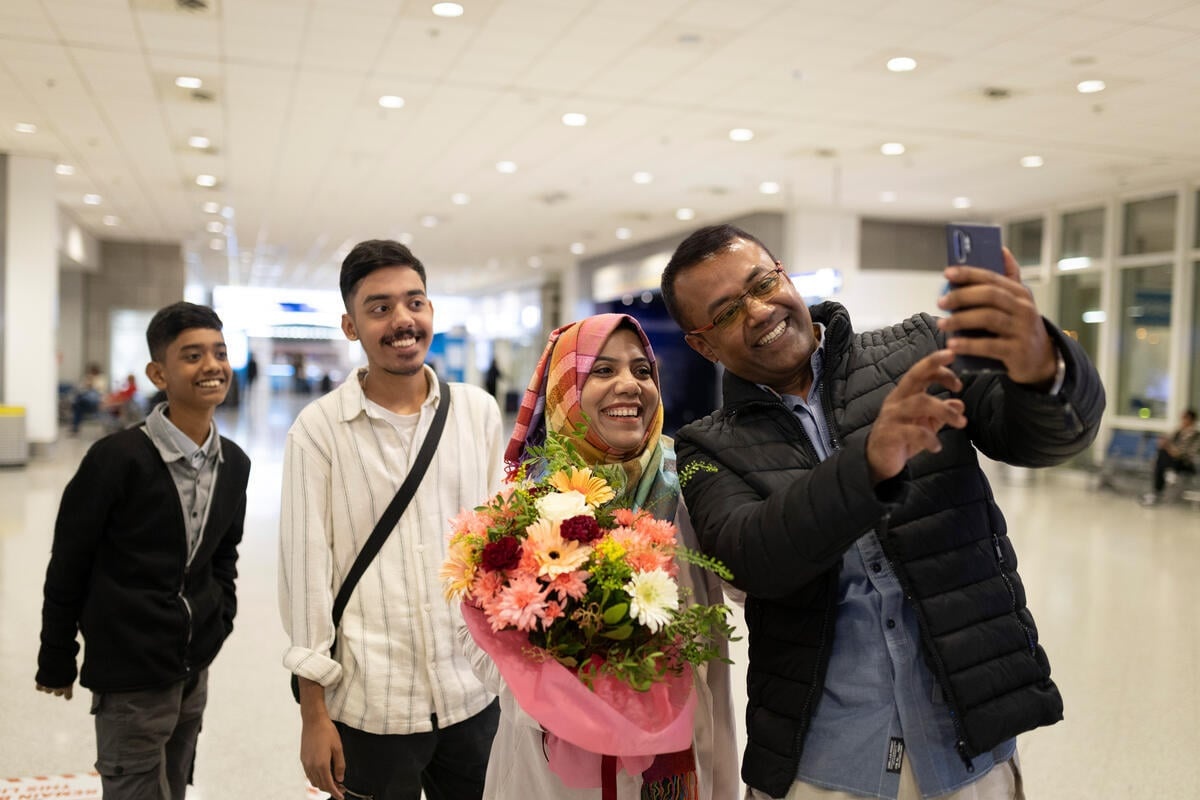 A man uses his phone to take a selfie with his wife and two sons at an airport.