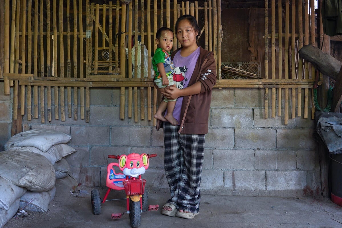 A woman carrying a baby boy stands in front of a cinder block and bamboo wall