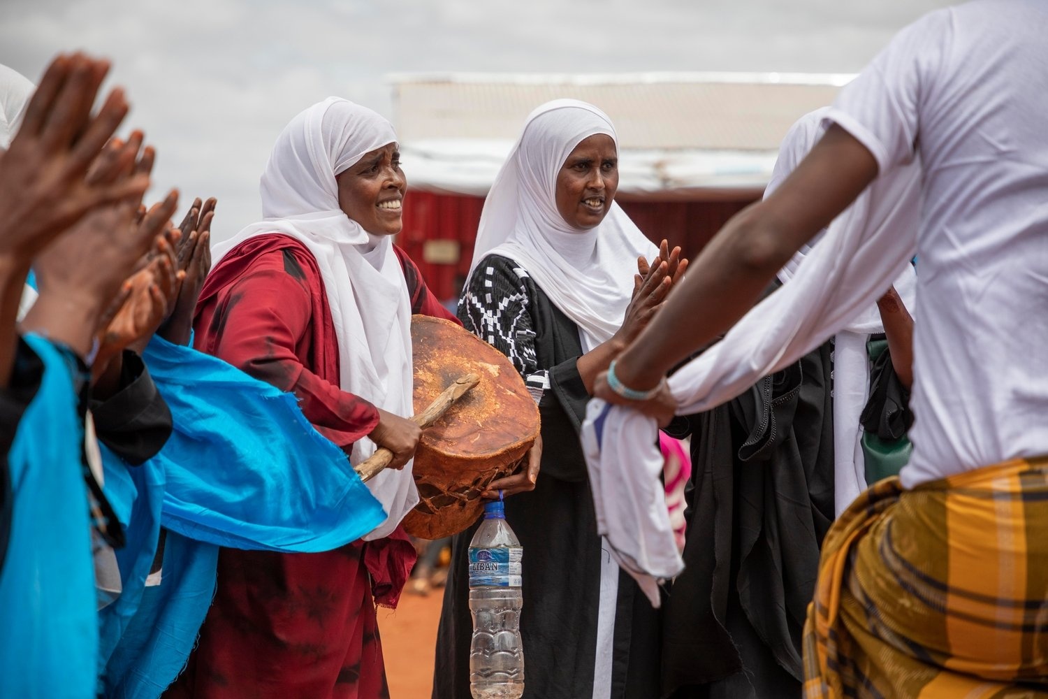 A woman beats a drum surrounded by people clapping and dancing.