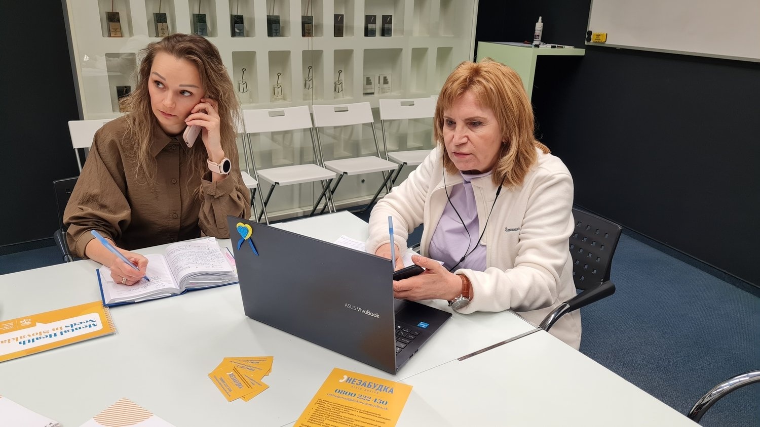 A woman wearing headphones looks at her laptop while a woman sitting next to her listens to her mobile phone while taking notes.