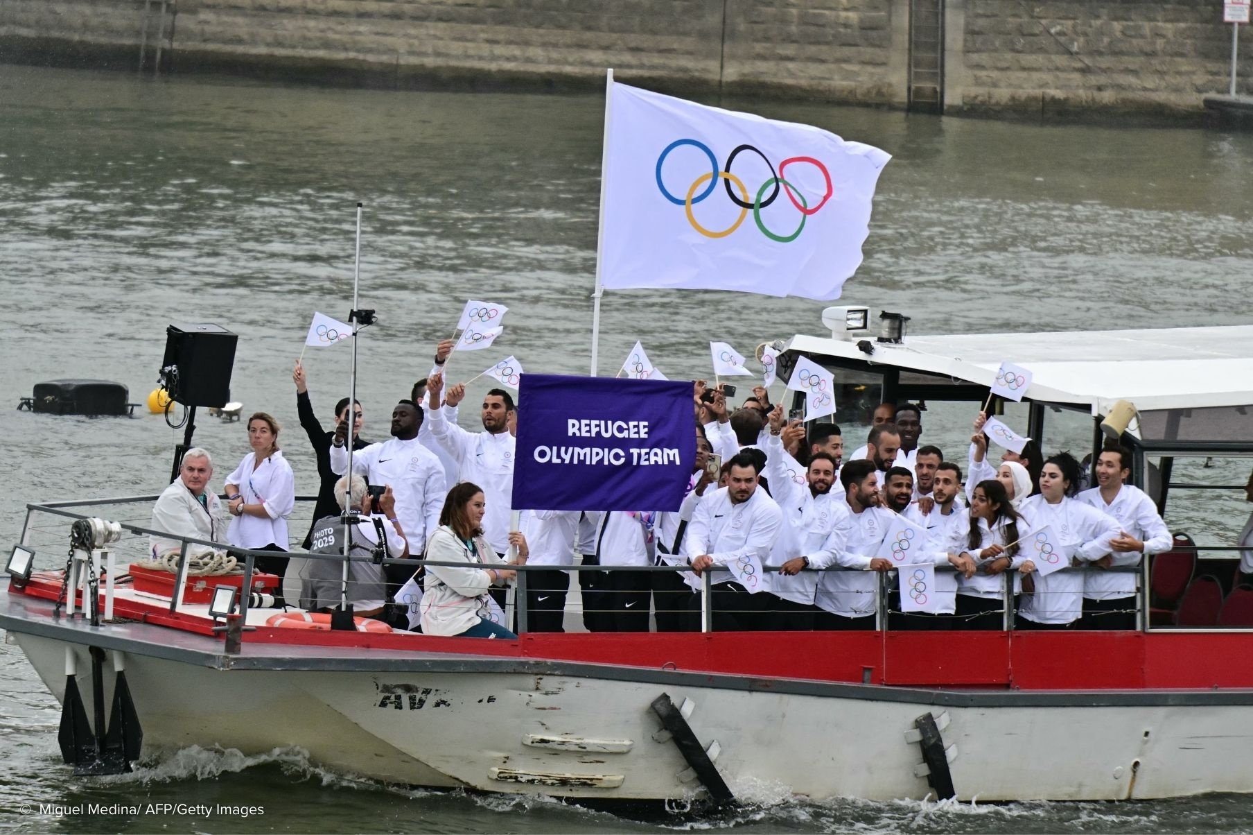 A group of people wearing white wave teams showing the Olympic rings on a boat going down a river.