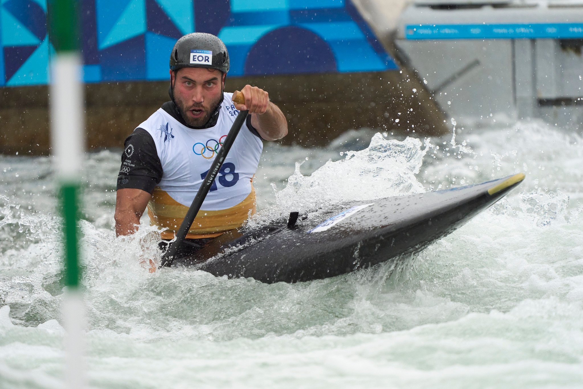Amir, wearing a helmet and Olympic bib, steers a canoe during a race.