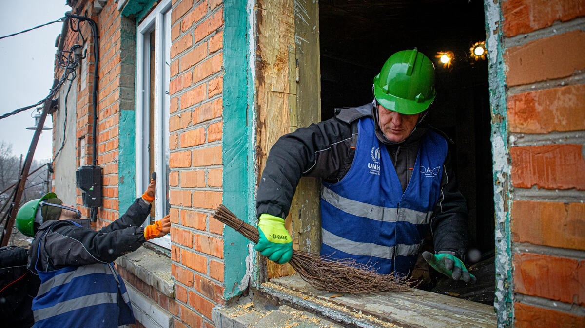 A workman in a green hard hat and blue vest sweeps a windowsill while behind him another worker installs a new window