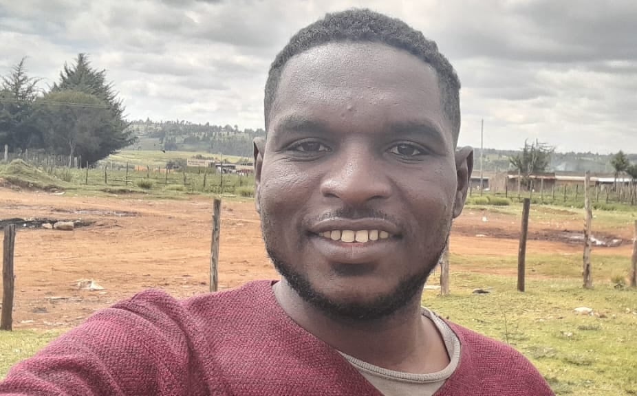 A selfie of a smiling man in front of a wire and wooden post fence, with a dirt road and countryside in the background