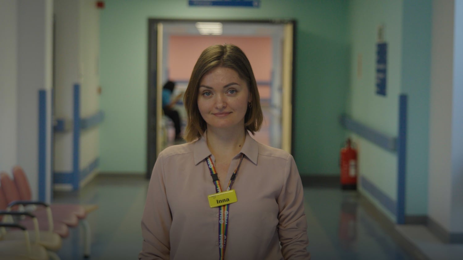 A woman stands in a hospital corridor wearing a yellow name tag reading 'Inna'