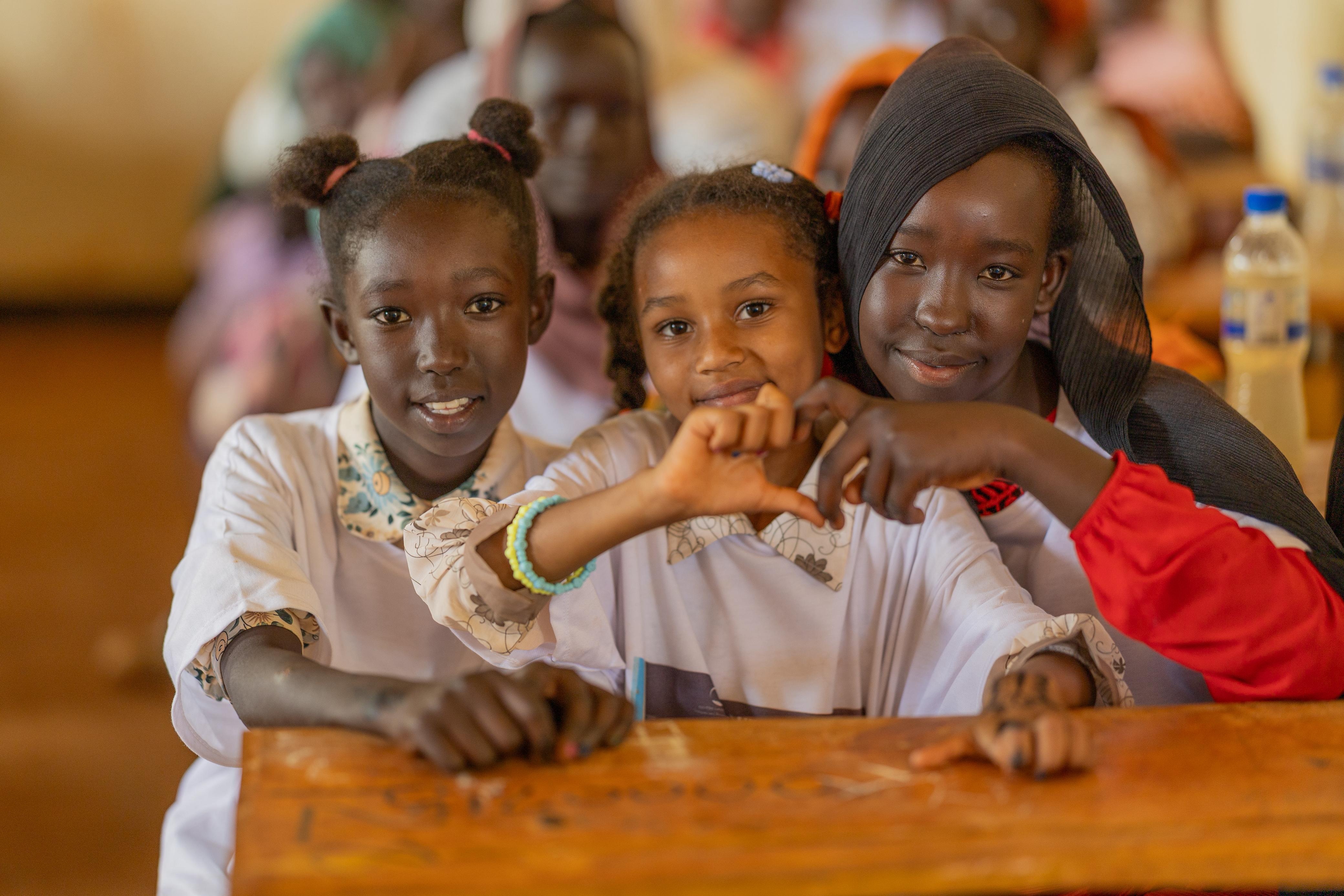 Three girls sit behind a desk in a classroom while two of them make a heart symbol with their hands