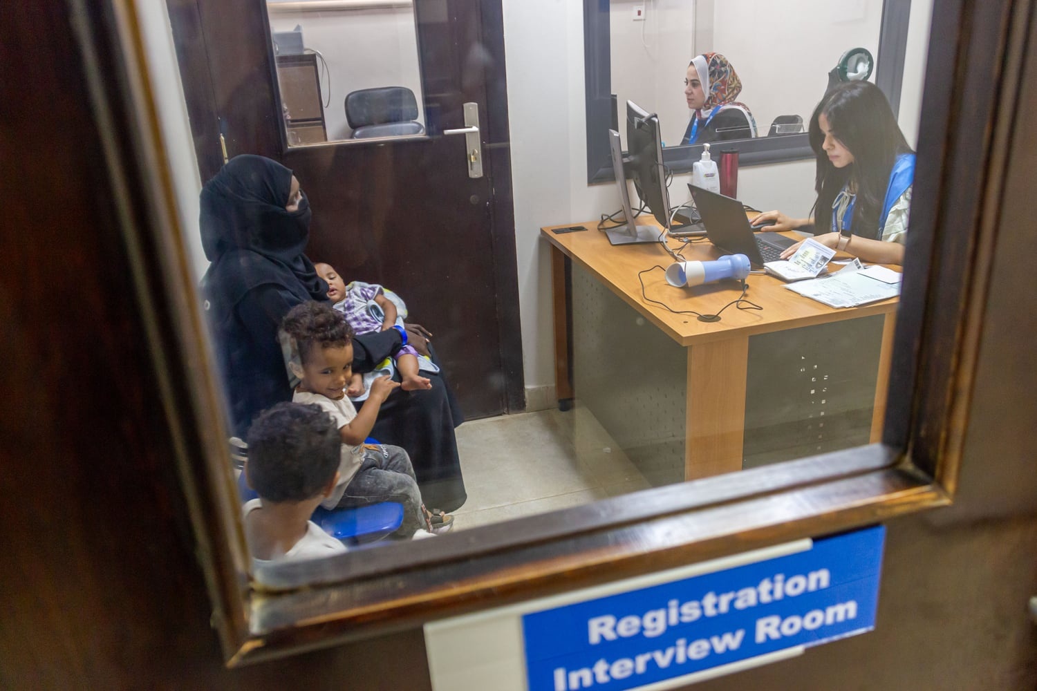 Through the window of a door labelled 'registration interview room', a woman and three children sits in front of a desk where a UNHCR staff member is typing on a computer 