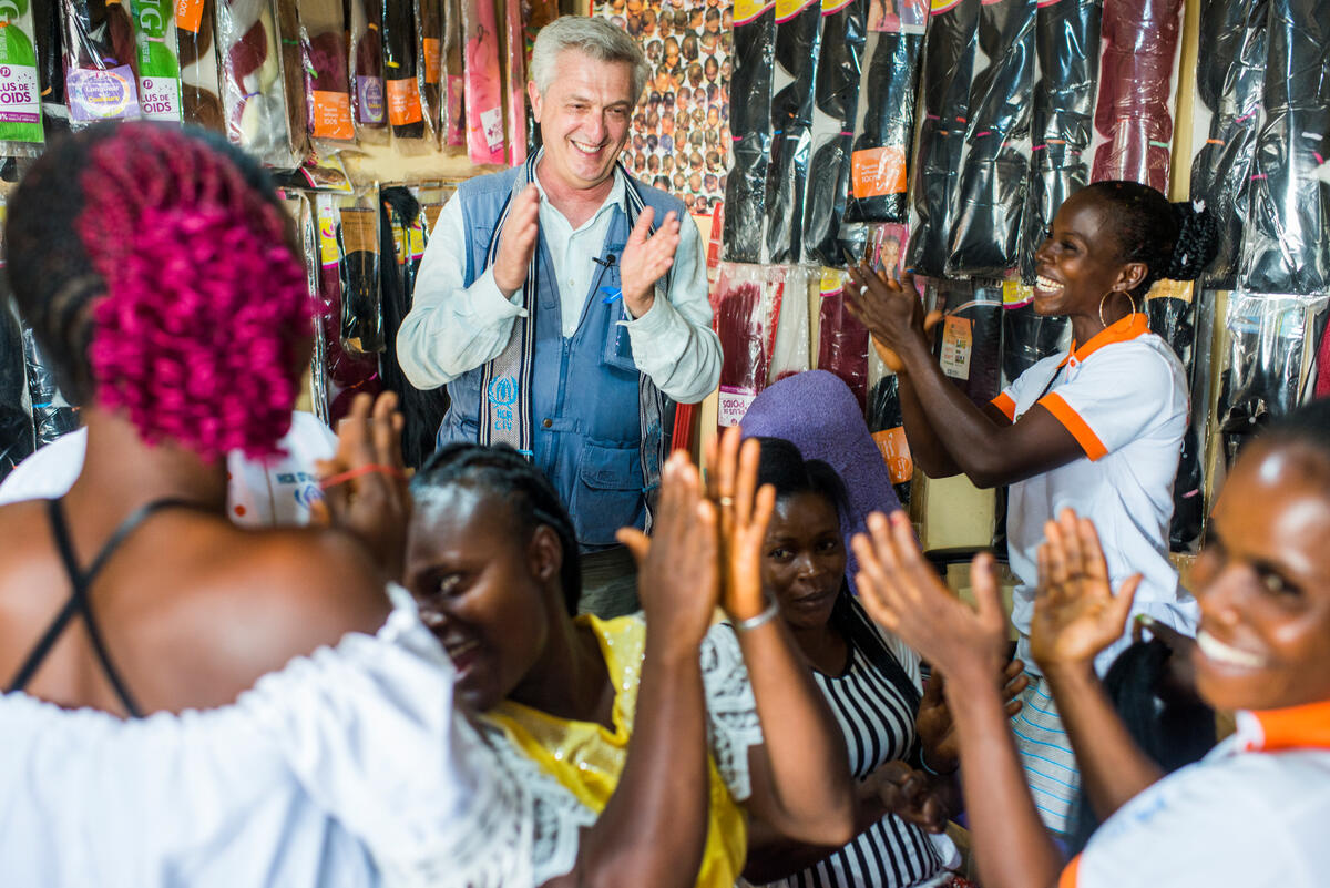 Filippo Grandi, UN High Commissioner for Refugees claps his hands in a hair salon, surrounded by cheerful, clapping Ivorian refugees.