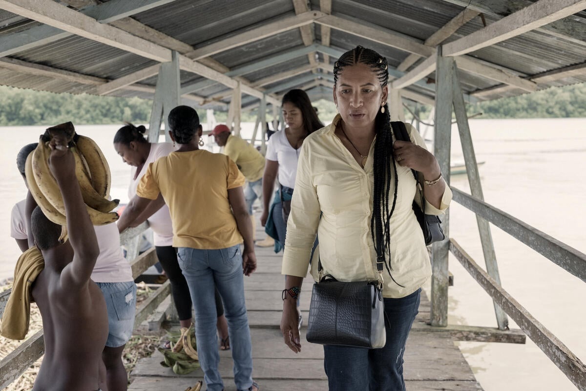 A woman walks past people on a covered wooden pier on a wide river.