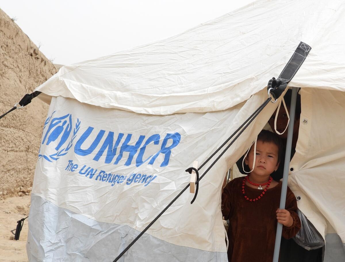 A child looks out from inside a white tent with a blue UNHCR logo 