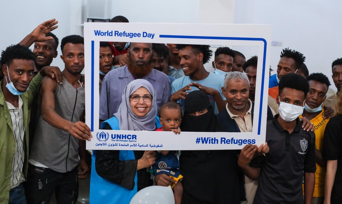 Refugees and UNHCR staff holding up a UNHCR frame which says 'World Refugee Day" and '#WithRefugees"