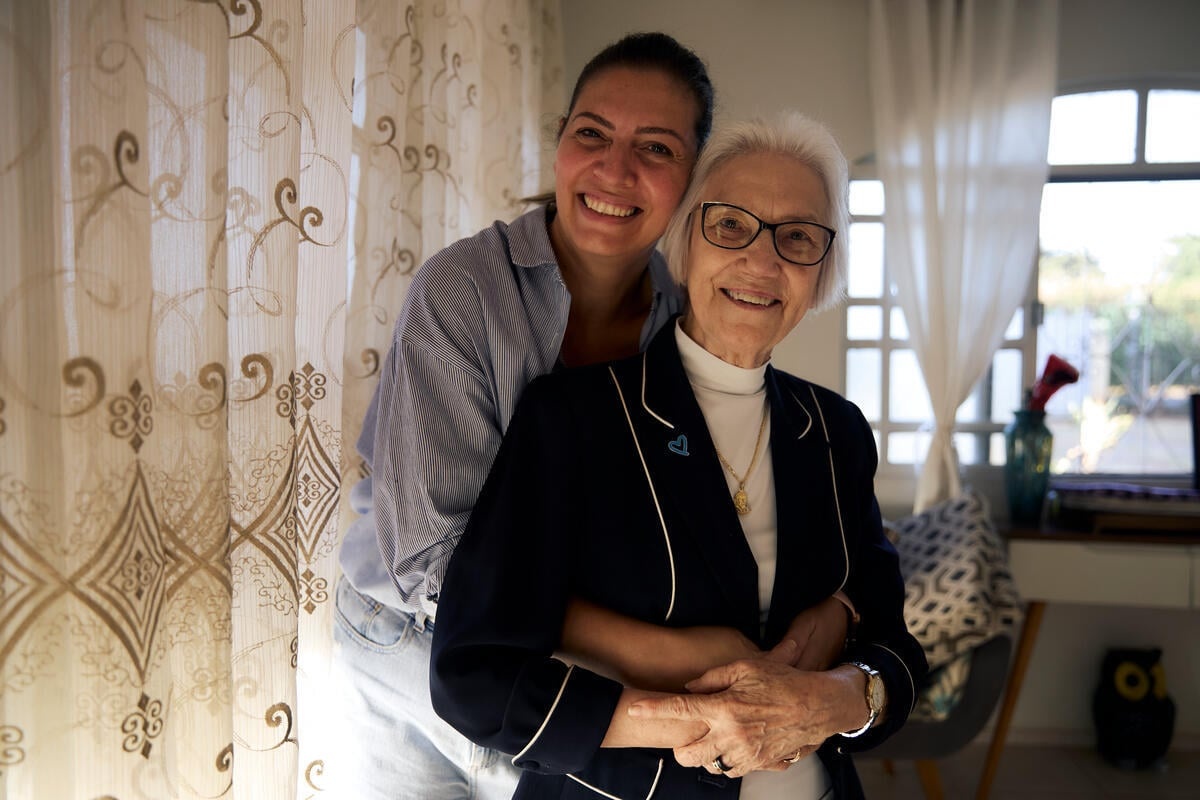 A smiling woman stands behind another woman with her arms around her next to a curtained window inside a house.