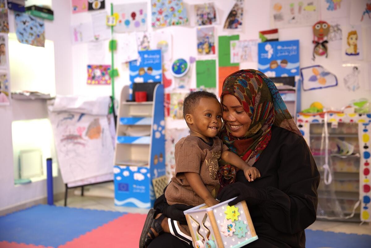 A smiling woman holds a toddler in a classroom decorated with children's artwork.