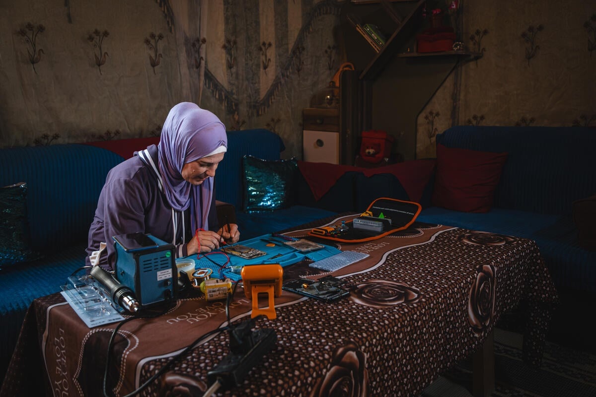 A woman sits at a table and uses tools to repair a phone.