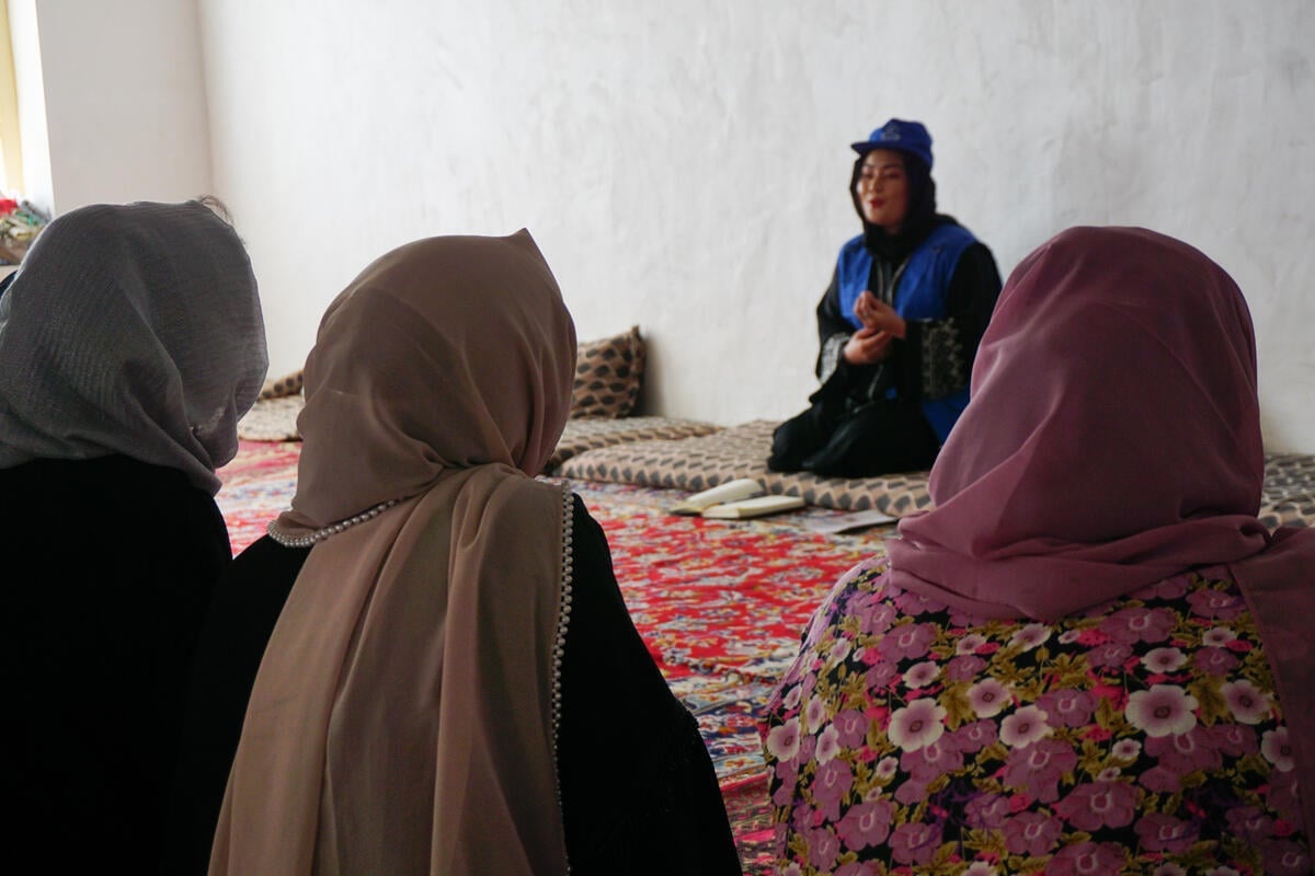 Three women with their back to the camera face another woman in a UNHCR blue hat and vest seated on a rug.
