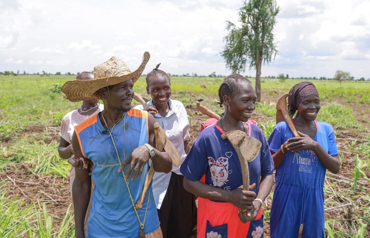 A man, a woman and three young people walk through a field smiling and holding hoes.