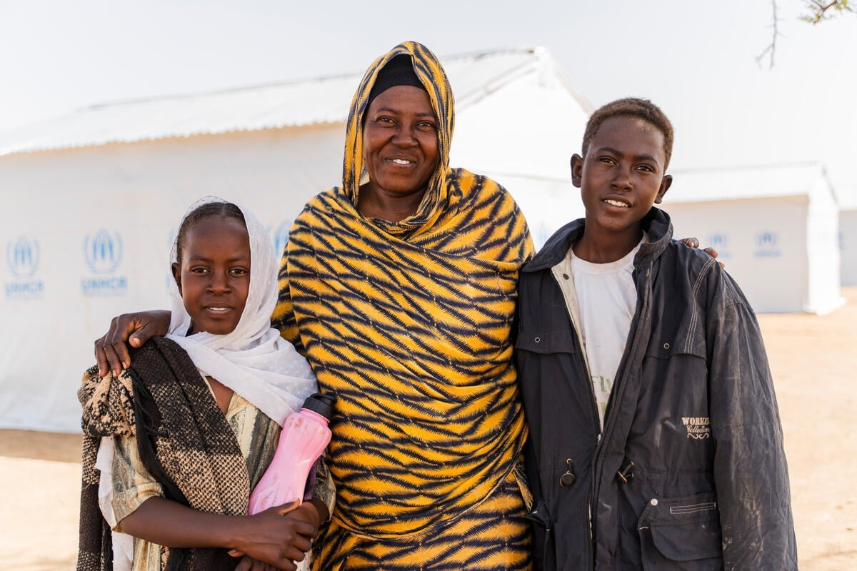 A mother stands with her young daughter and son in front of their shelter