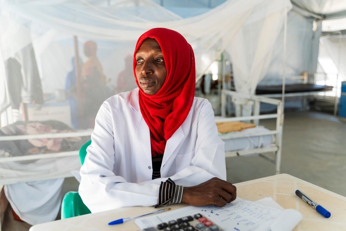 A female doctor wearing white coat and red hijab sits down at a desk in a hospital