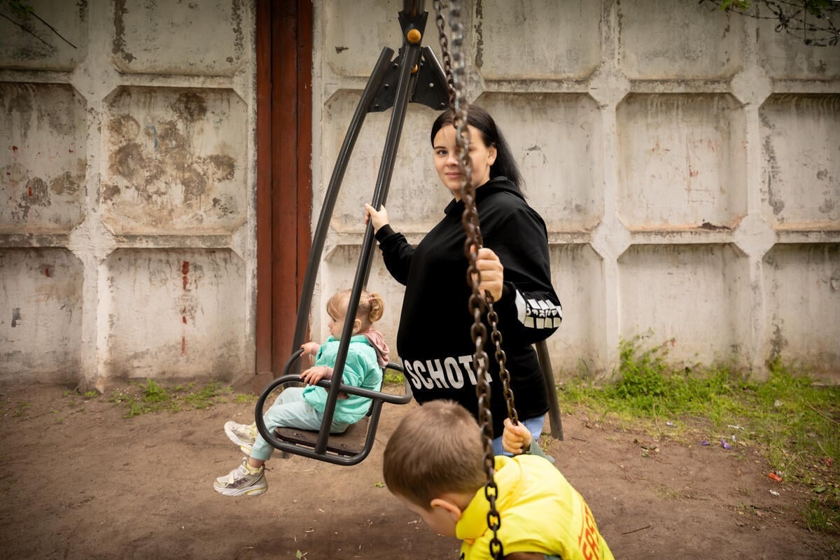 A pregnant mother pushes her two young children on park swings