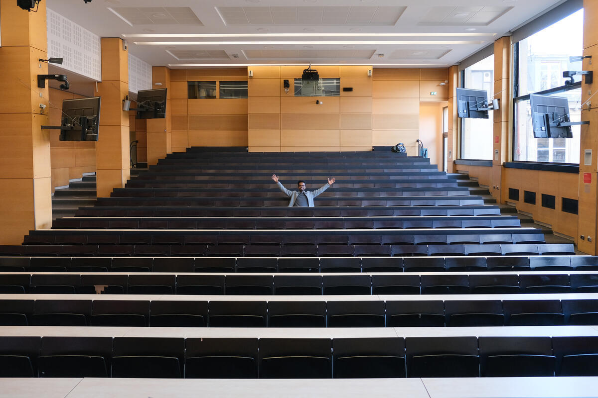A smiling man raises both arms while seated in the middle of an empty university lecture hall.