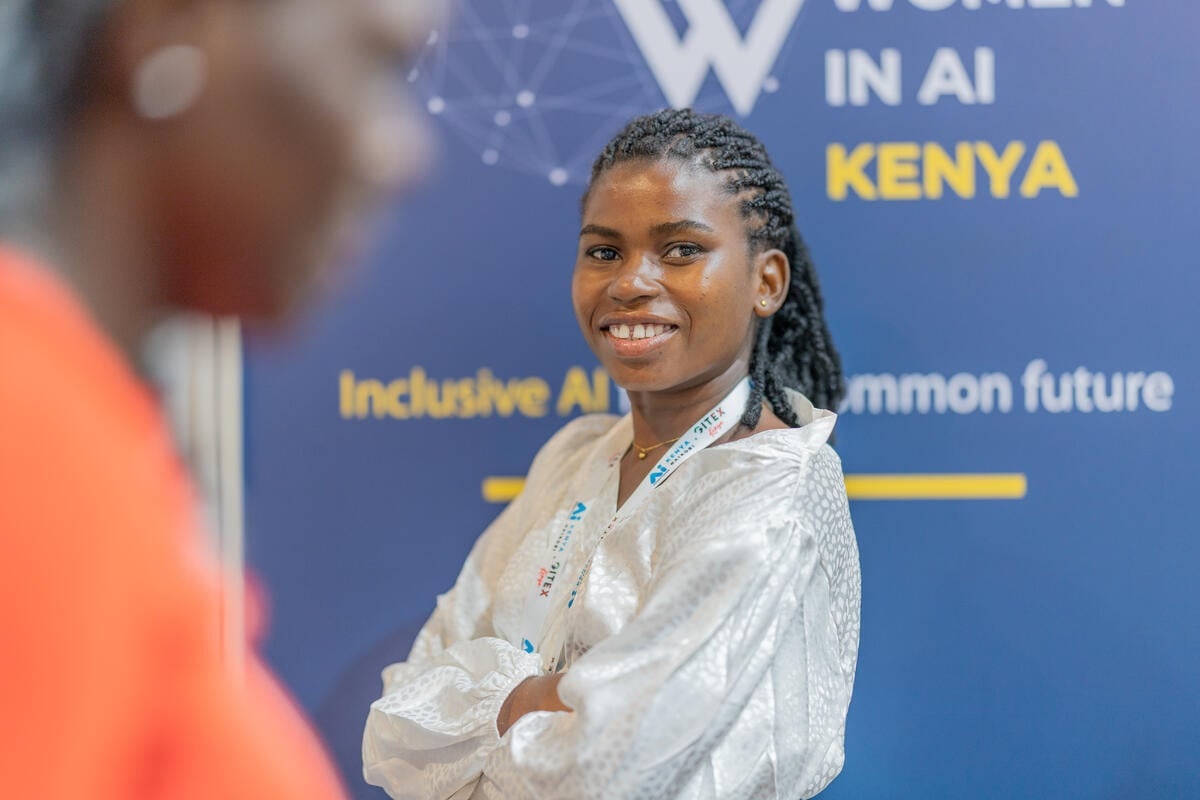 A smiling young woman with crossed arms stands in front of a conference poster for an event on women in AI in Kenya
