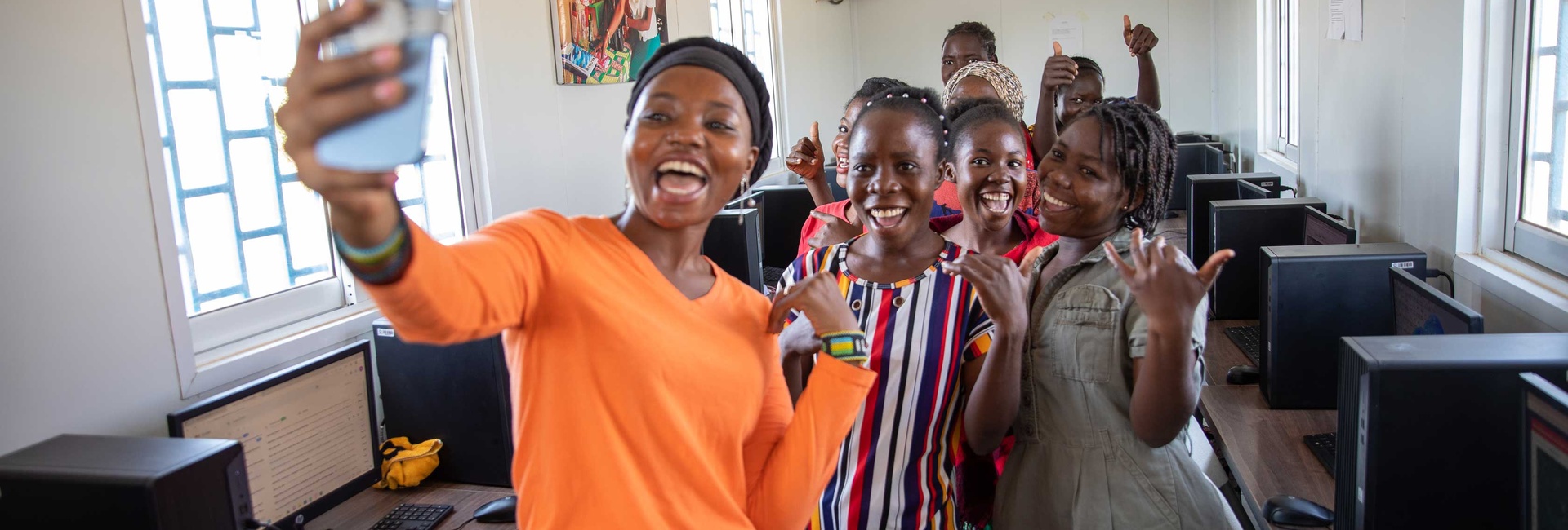 A woman in a bright orange shirt takes a selfie with a group of young women in a computer lab.