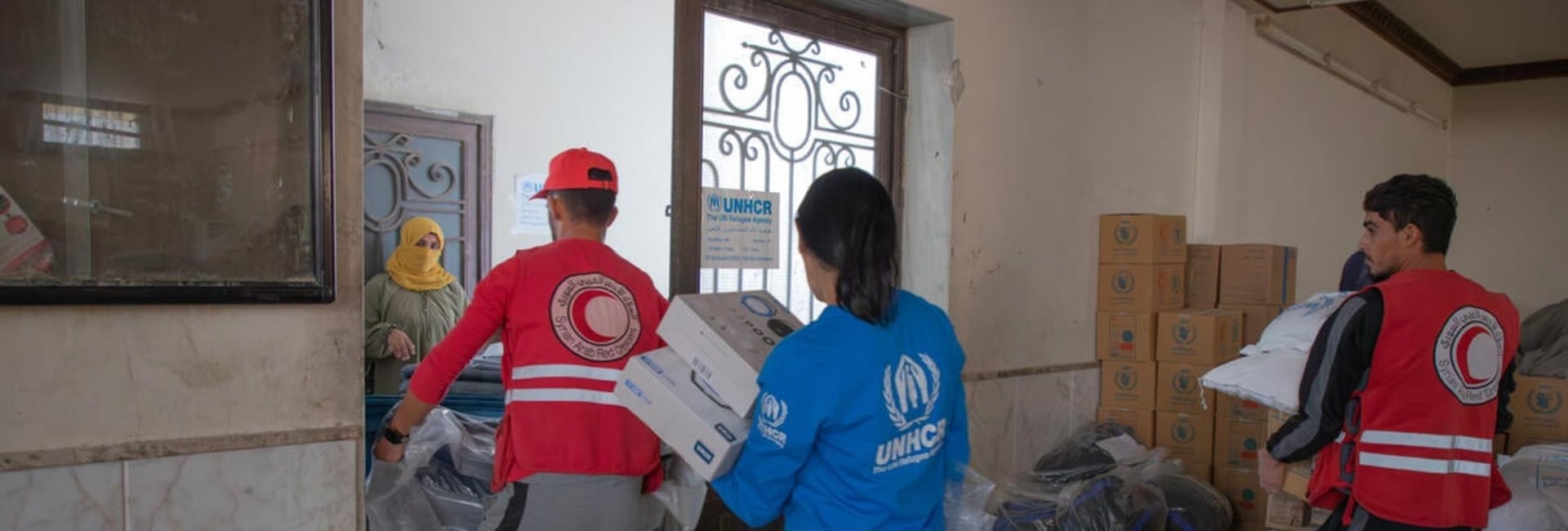 UNHCR and Syrian Arab Red Crescent staff carry boxes and bags of supplies out of a room.