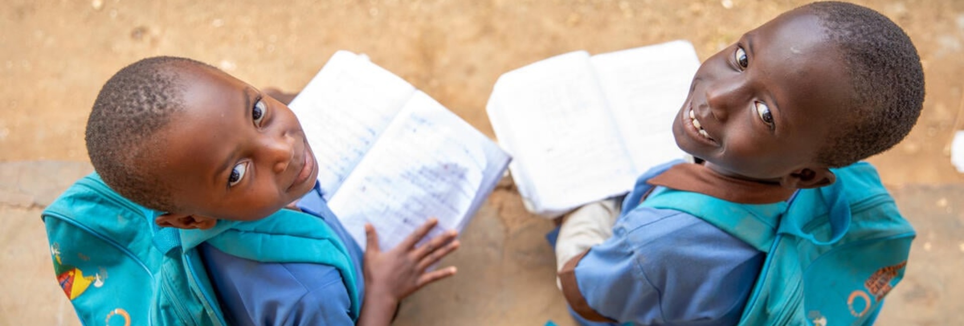 Two girls, 7- and 8-years-old, from Central African Republic smile up at the camera from where they're sitting outside, wearing matching 'Education cannot wait' backpacks and holding open books.