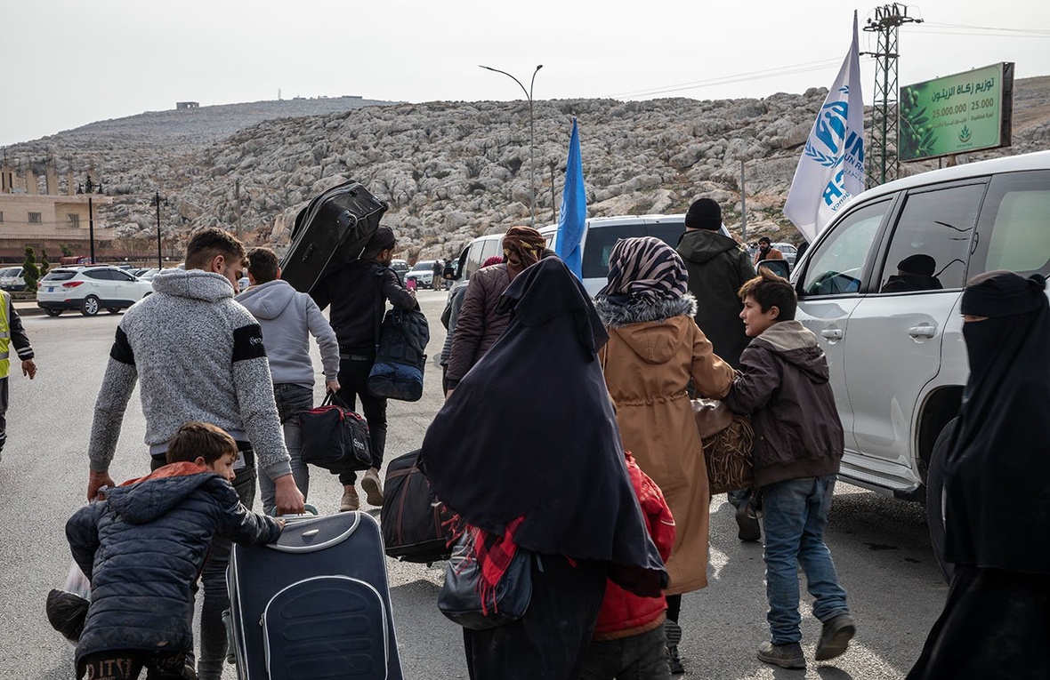 A group of people walk on a road with their belongings, towards a large rocky outcrop