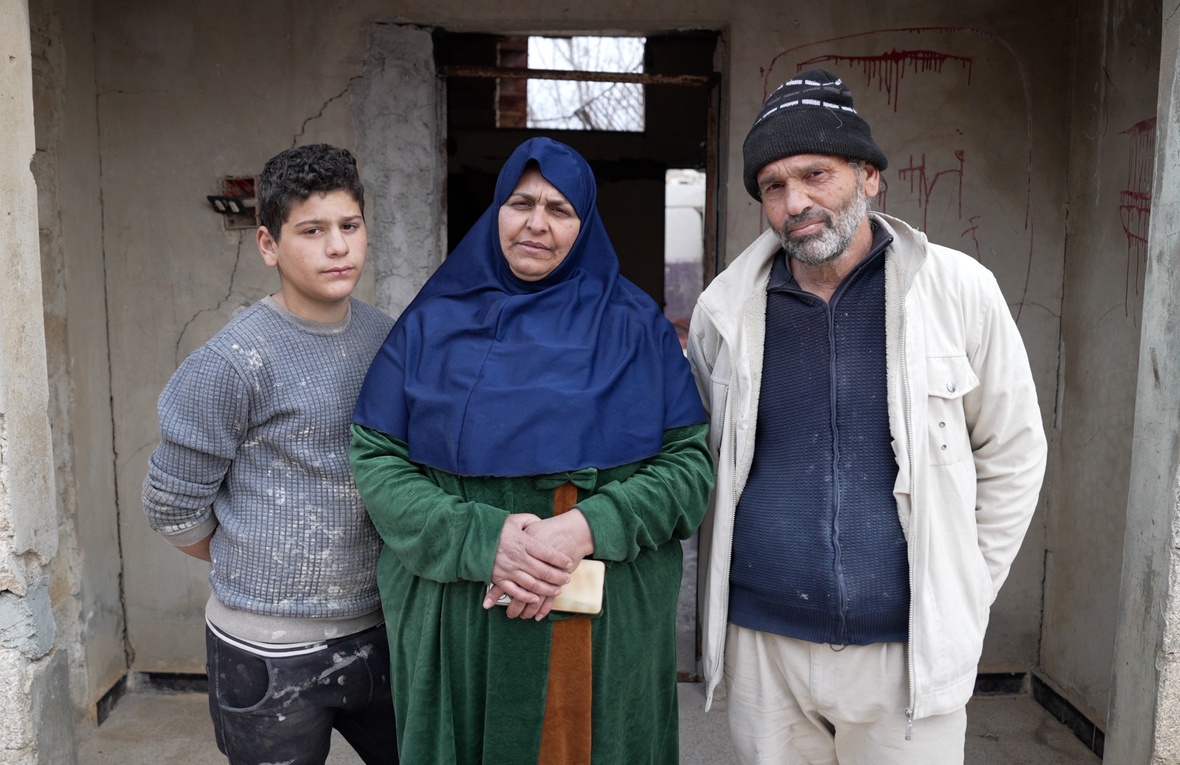 A couple and their teenage son stand outside a damaged house with graffiti on the wall