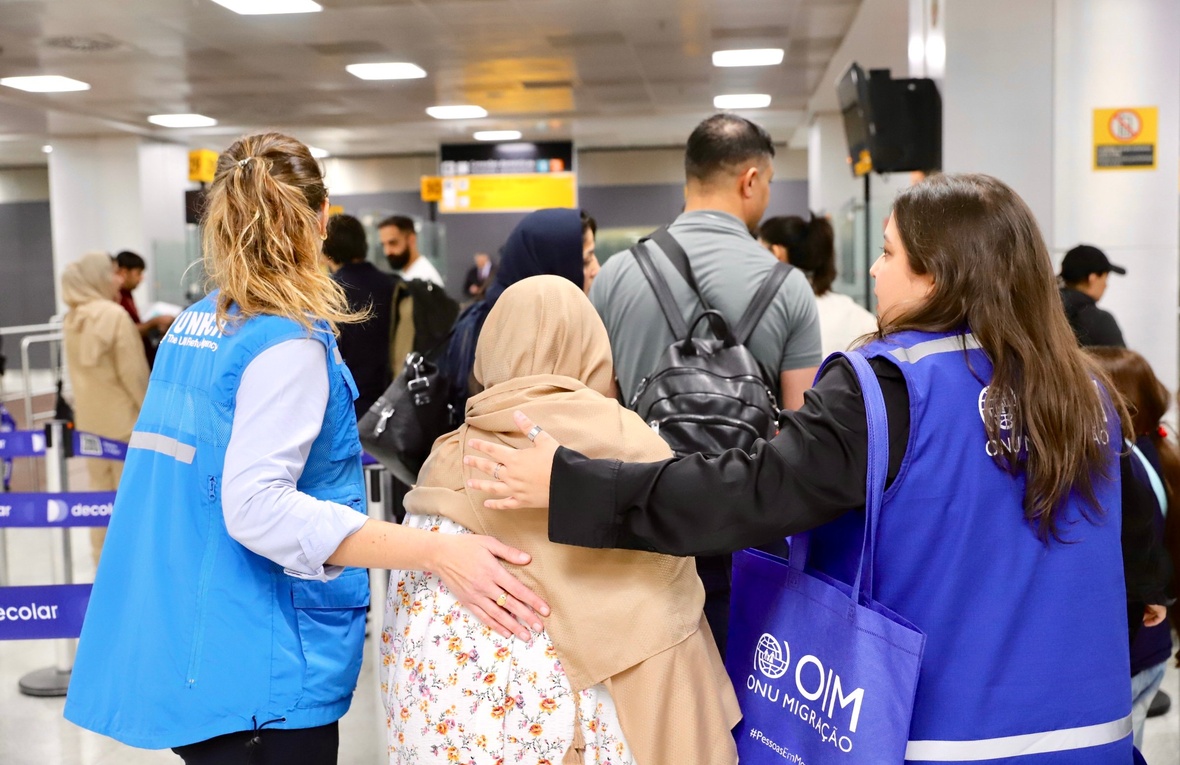 Staff from UNHCR and IOM walk through a crowded airport with their arms around a woman