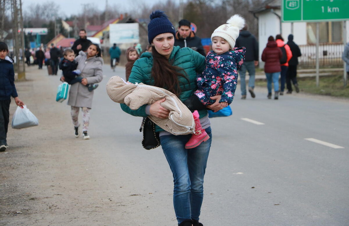 A young woman walks along the side of a road carrying a toddler and a baby in her arms 