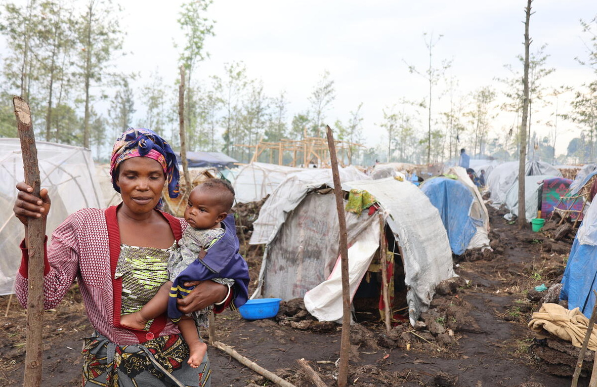 A woman holding a baby stands in front of temporary shelters in a displacement camp in DR Congo.